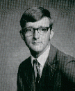 A black and white portrait of a young man wearing glasses and a suit, with a neutral expression, set against a plain background.