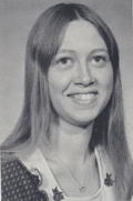 A black and white portrait of a young woman with long hair, smiling at the camera, wearing a light-colored top with decorative elements.
