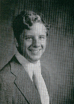 A black and white portrait of a young man with curly hair, wearing a suit and tie, smiling at the camera.