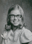 A young girl with long, wavy hair and glasses smiles at the camera, wearing a light-colored blouse with a collar.
