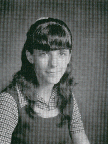 A black and white portrait of a young woman with long hair and bangs, wearing a patterned blouse and a dark dress, looking directly at the camera.