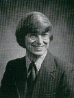 A black and white portrait of a smiling man with long hair and glasses, wearing a suit and tie.