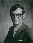 A black and white portrait of a man with glasses, wearing a suit and tie, smiling at the camera.