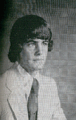 A young man with long hair wearing a light-colored suit and tie, posed against a textured background.