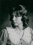 A black and white portrait of a young woman with wavy hair, wearing a checkered dress with a white collar, gazing to the side.