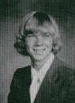 A black and white portrait of a young man with long, wavy hair, smiling while wearing a suit and tie.