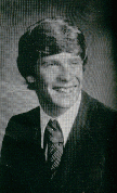 A black and white portrait of a young man with a hairstyle typical of the 1970s, wearing a suit and tie, smiling at the camera.