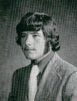 A black and white portrait of a young man with long, wavy hair and a mustache, wearing a suit and tie, looking slightly to the side.