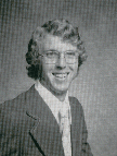 A black and white portrait of a smiling man with curly hair, wearing glasses and a suit, posed against a plain background.