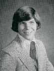 A man with a distinctive hairstyle and a patterned suit smiles at the camera in a studio portrait.
