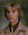 A young man with light blonde hair, wearing a beige suit and tie, poses for a formal portrait against a blurred background.
