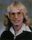 A young man with long, wavy blonde hair and glasses, wearing a black jacket over a white shirt with a collar, smiles at the camera in a school portrait.