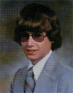 A young man with long, wavy hair and sunglasses, wearing a light gray suit and a striped tie, poses for a portrait against a blurred background.