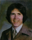 A young man with dark, wavy hair and a broad smile, wearing a brown suit and patterned tie, poses for a portrait against a blurred background.