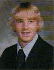 A young man with blonde, wavy hair wearing a dark suit and tie, smiling at the camera in a formal portrait.
