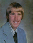 A young man with a bowl haircut, wearing a light blue suit and a checkered shirt, smiles at the camera in a studio portrait.
