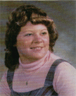 A young woman with curly hair, wearing a pink turtleneck and a denim overall, smiles softly against a blurred background.