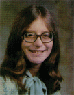 A young woman with long brown hair and glasses smiles at the camera, wearing a light blue blouse with a bow tie.