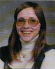 A smiling young woman with long brown hair, wearing round sunglasses and a turtleneck sweater, poses for a photo against a neutral background.
