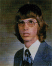 A young man with long hair and glasses, wearing a suit and tie, poses for a formal portrait.