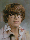 A young person with curly hair and large glasses, wearing a patterned shirt, poses for a school portrait.