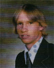 A young man with long, light brown hair wearing a dark suit and a patterned tie, posing for a portrait against a blurred background.