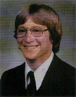 A young man with glasses and medium-length hair, smiling while wearing a suit and tie against a blurred background.