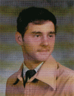 A young man with dark hair and a mustache, wearing a light-colored coat and a tie, poses against a softly blurred background.