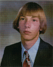 A young man with a bowl haircut, wearing a suit and tie, poses for a formal portrait against a blurred background.