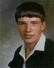 A young man with short hair and a slight smile, wearing a black jacket over a white shirt, posed for a school portrait against a blurred background.