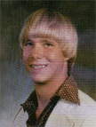 A young man with a bowl haircut and a big smile, wearing a white suit jacket over a patterned shirt, posed against a blurred background.
