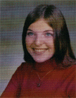 A young woman with long brown hair and blue eyes smiles while wearing a red turtleneck sweater, seated against a blurred background.