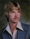 A young man with a mustache and shoulder-length hair, wearing a blue suit with a white collar, poses for a portrait against a blurred background.