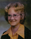 A young woman with curly blonde hair and glasses smiles for a portrait, wearing a black jacket over a yellow collared shirt.