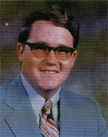 A man with glasses and a light blue suit, smiling at the camera, with a patterned tie and a blurred background.