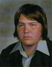 A young man with long, dark hair and a serious expression, wearing a collared shirt and a jacket, posed for a portrait against a muted background.