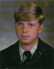 A young man with light brown hair styled in a side part, wearing a dark suit and tie, looking directly at the camera with a neutral expression.