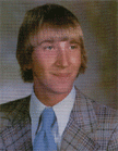 A young man with a bowl haircut, wearing a plaid suit and a light blue tie, smiles for the camera against a blurred background.