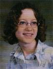 A young girl with curly hair and glasses smiles for a portrait, wearing a patterned shirt.