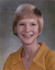 A smiling young woman with short blonde hair, wearing a yellow blouse, poses for a portrait against a soft, blurred background.