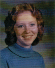 A young woman with curly red hair smiles at the camera, wearing a blue turtleneck sweater against a softly blurred background.