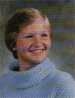 A smiling young woman with short hair wearing a light blue turtleneck sweater, set against a blurred background.