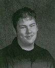 A young man with short, dark hair and a slight smile, wearing a black shirt, is posed against a textured gray background.