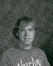 A black and white portrait of a young man with short, wavy hair, wearing a graphic t-shirt, looking directly at the camera.