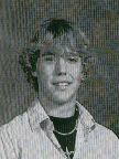 A black and white portrait of a young man with curly hair, wearing a collared shirt and a necklace, smiling at the camera.