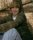 A young man with shaggy hair is sitting against a tree in a wooded area, smiling at the camera, wearing a green striped sweater and light blue jeans.