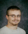A young man with short hair and glasses smiles at the camera, wearing a light-colored shirt against a dark background.