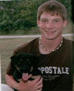 A young man with short brown hair smiles while holding a black puppy in his arms, set against a grassy background.