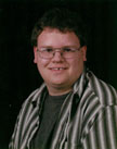 A young man with glasses and a striped shirt smiles at the camera against a dark background.