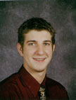 A young man with short, styled hair and a friendly smile, wearing a maroon shirt and patterned tie, poses against a gray backdrop.